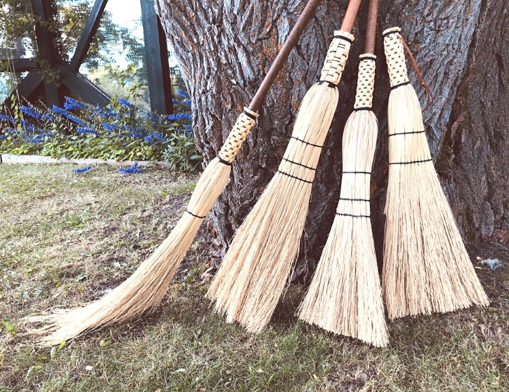 photo of four handmade brooms propped up against a tree trunk with autumn foliage in the background